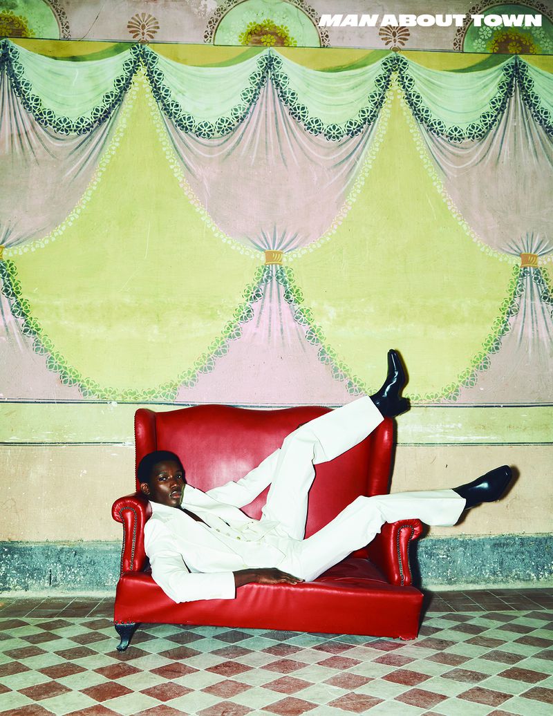 An adult male in a white suit and black shoes reclines in a red chair against a decorative wall in this high-fashion, full shot.