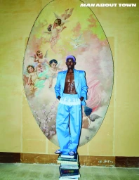 Model Ibra Niakate stands on a stack of books wearing an open light blue suit against an oval cherub painting for Man About Town.