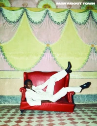 Model wearing a white suit lying sideways on a bright red sofa against an ornate green and pink patterned wall