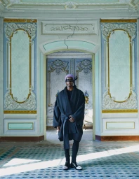 Model Ibra Niakate wearing an oversized belted dark robe coat and eye covering in an ornate, frescoed historic room.