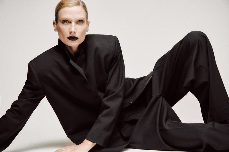 Blond adult female model in a black suit lying down on a bright white background. Bold black lipstick. Studio lighting. Fashion shot.