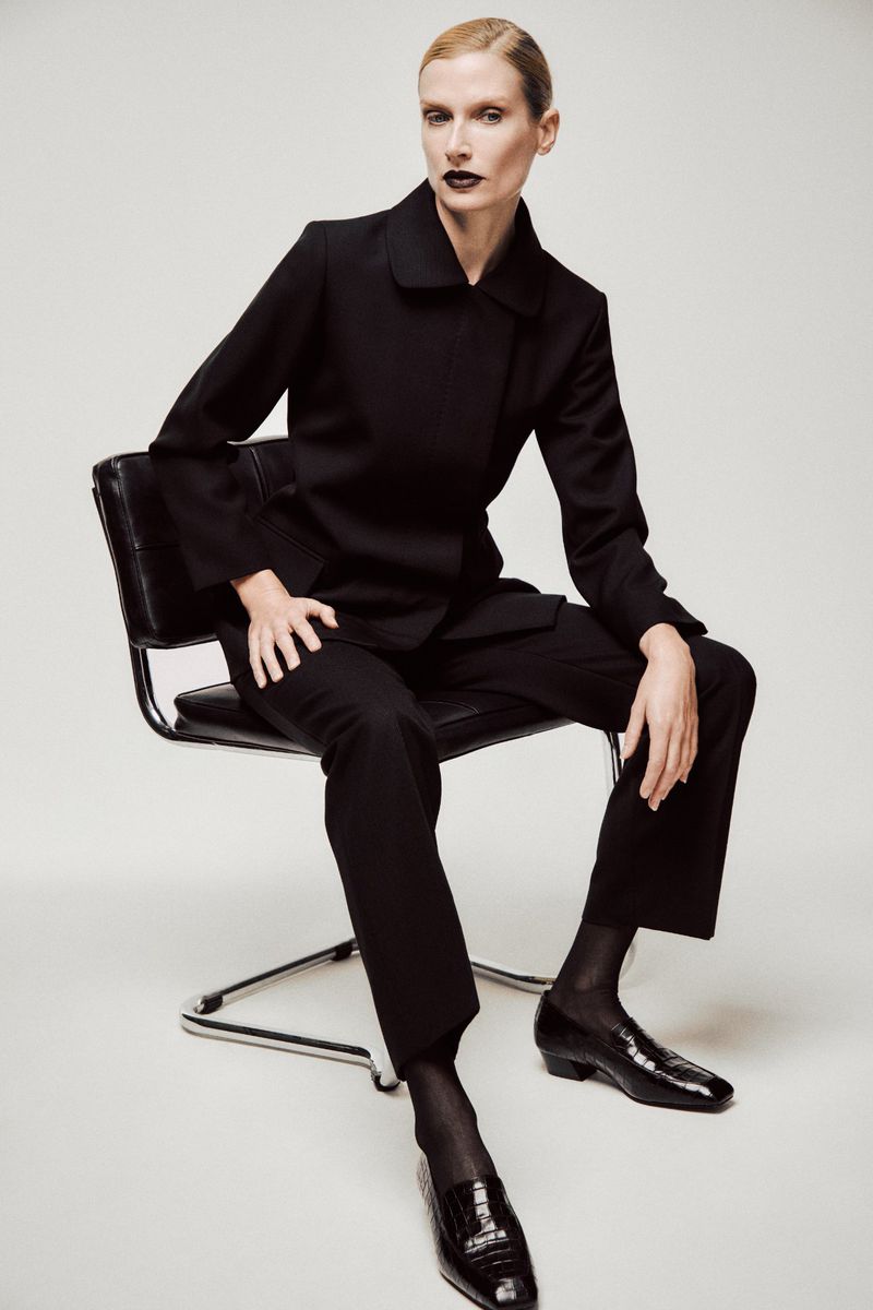 Fashionable woman in black suit and stockings sitting on a metal and leather chair in a photo studio, minimalist background, bold lipstick.