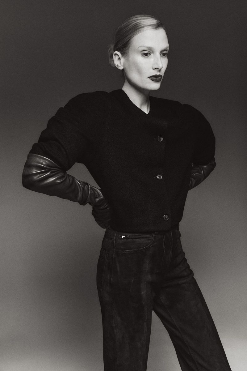 Monochrome portrait of a woman in a black jacket with leather sleeves and black trousers, hands on hips, against a grey backdrop.