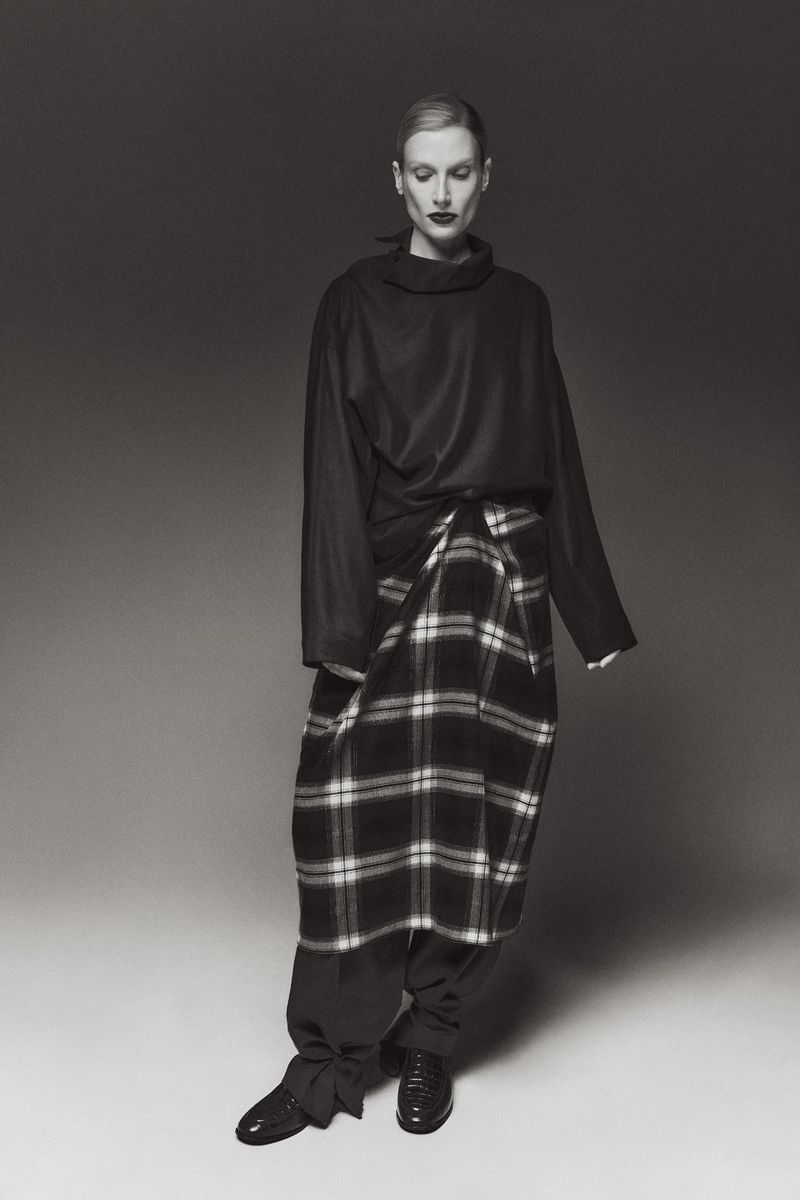Elegant woman in a fashion shot wearing a black turtleneck sweater, plaid skirt-pants, and black shoes in a studio setting on gray background.
