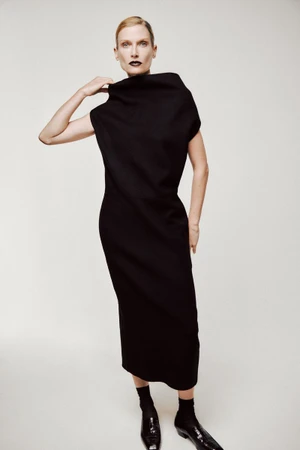 Model Nina Heimlich poses in a sculptural high-neck black dress, black socks, and loafers against an off-white seamless backdrop.