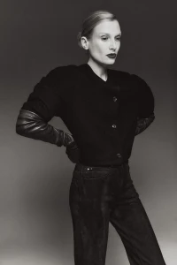 Black and white portrait of a woman in a dark buttoned jacket and dark trousers standing against a gradient studio backdrop.