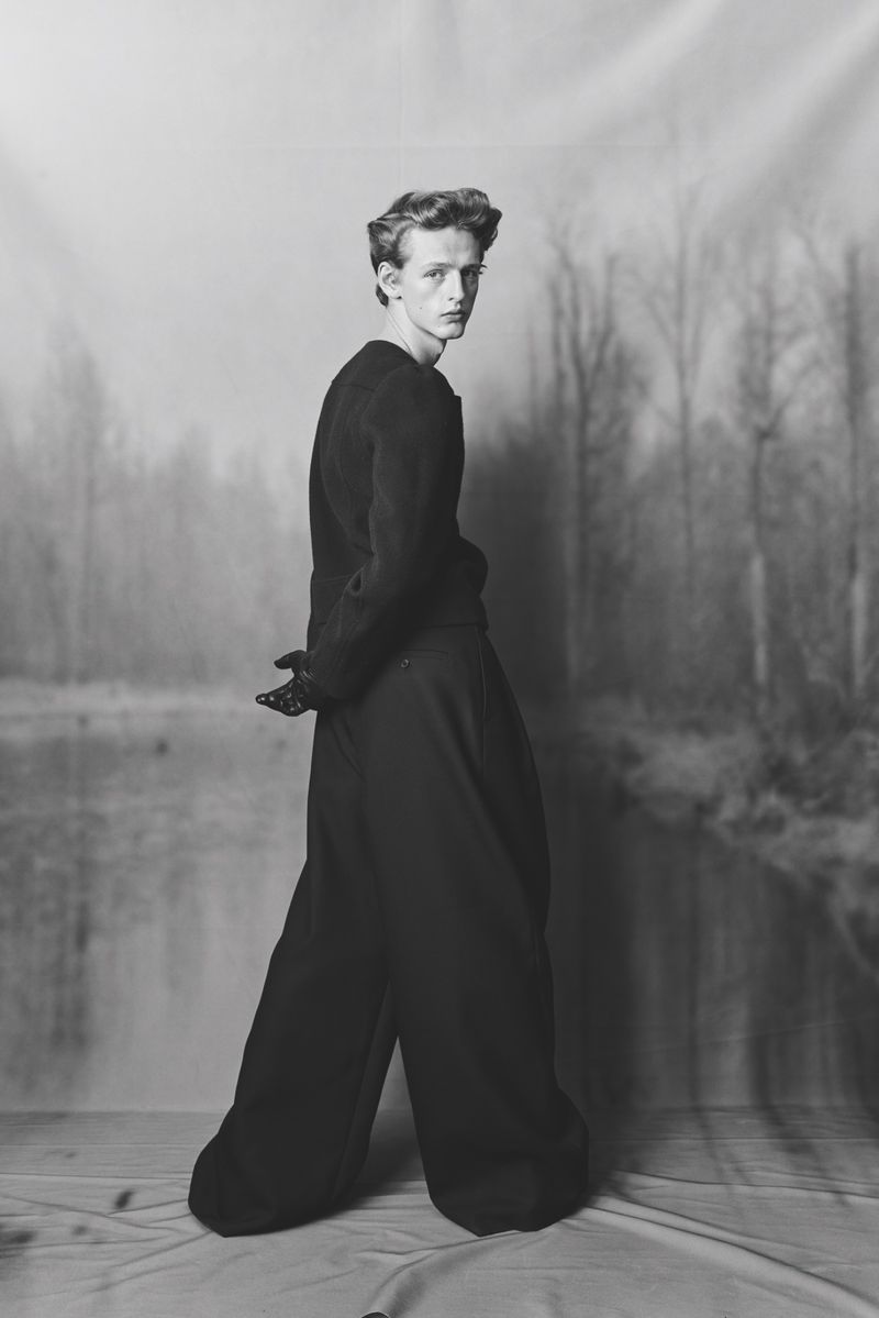 Monochrome portrait of a slender young adult in a dark sweater and voluminous pants, looking over their shoulder in an outdoor setting.