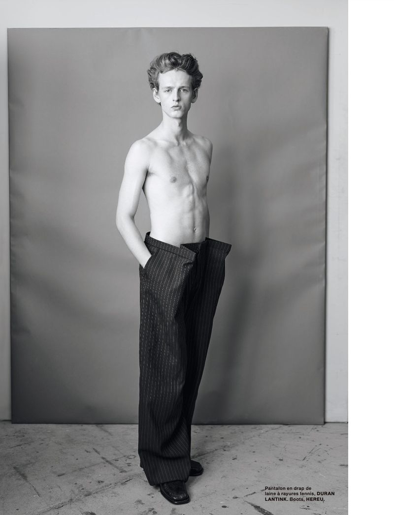 Black and white portrait of a young adult male posing in a photo studio setting wearing only large pinstripe pants.