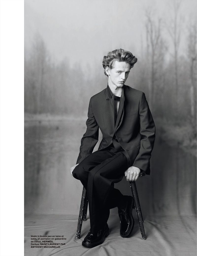 Monochrome portrait of a young man seated on a stool wearing a tailored black suit and dress shoes against a misty forest backdrop.