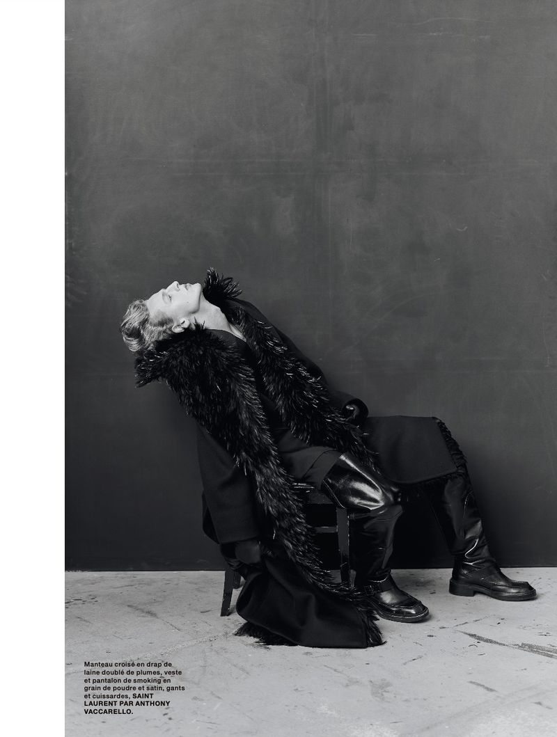 Fashion editorial shot of a woman in a feathered shawl, coat, thigh-high boots, and a tilted head pose against a gray backdrop.