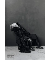Model leaning back in a chair wearing a voluminous black feathered coat and tall leather boots against a grey studio backdrop.
