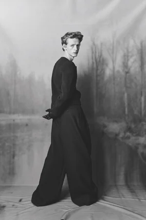 Monochrome fashion editorial portrait of a young man in dark trousers and sweater standing before a painted forest backdrop