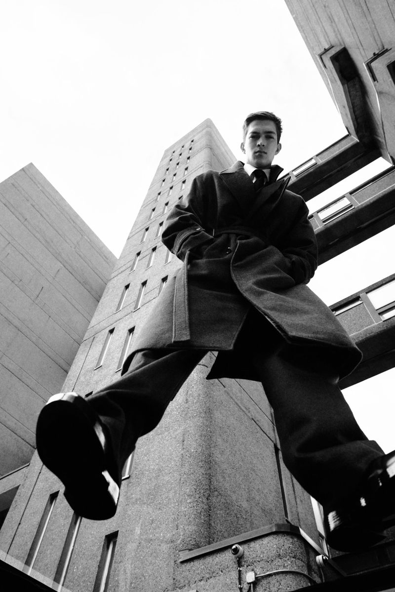 Fashion shot of a young man wearing a coat and dress pants in front of concrete buildings, captured from a low angle in black and white.
