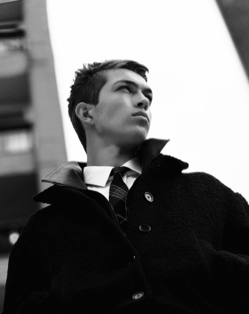 Portrait of a young adult man in a black coat and tie, looking upward with a serious expression, photographed in black and white against a building.