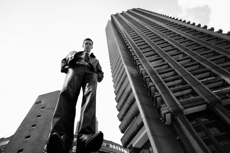 Low-angle shot of a stylish adult male in a leather jacket and wide-leg pants standing in an urban setting with high-rise buildings.