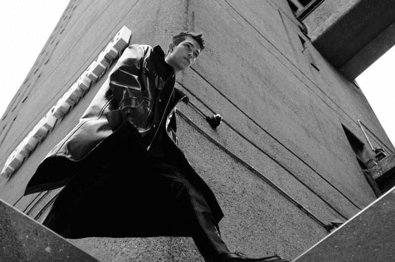 Low-angle black and white shot of a young adult man in a leather jacket and dark pants against a concrete building, looking up.