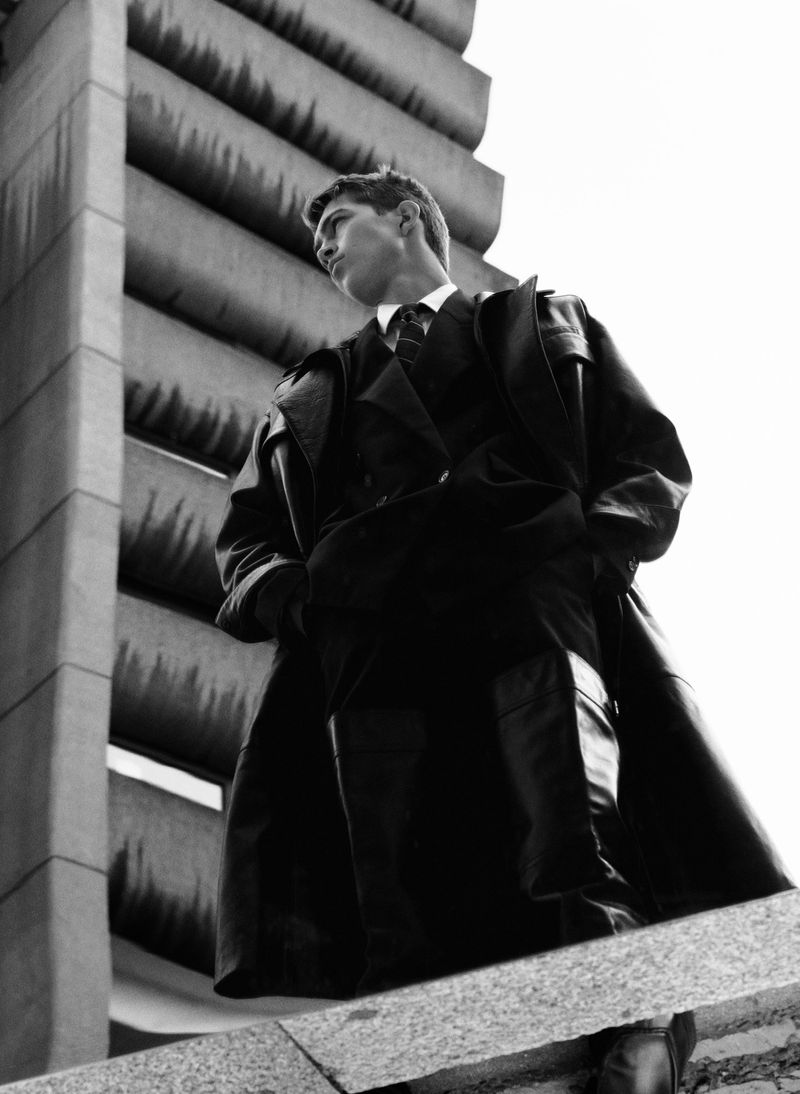 Monochrome low-angle shot of a male model in a trench coat and suit, posing against a brutalist concrete building on a cloudy day.