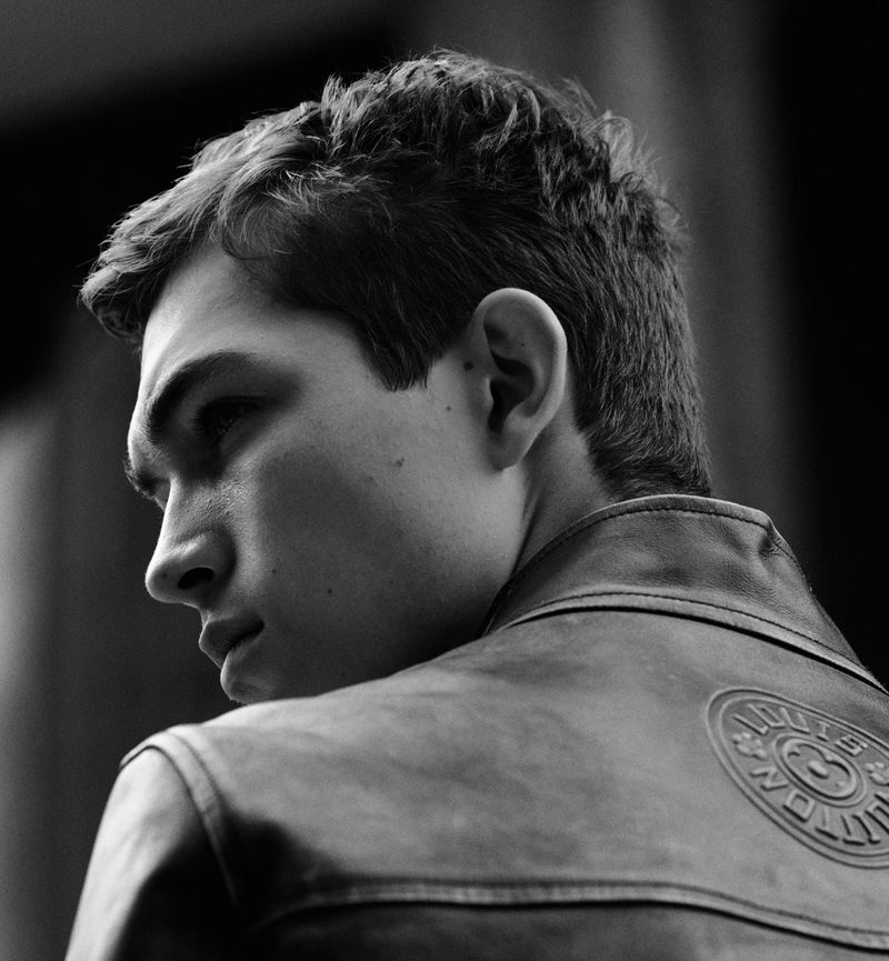 Monochrome portrait of a young adult male in a leather jacket with a logo over his left shoulder, facing away from the camera.