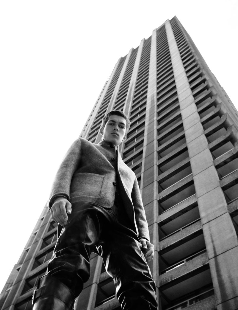 Man in a wool coat and leather pants stands in front of a concrete building, shot from a low angle in black and white.