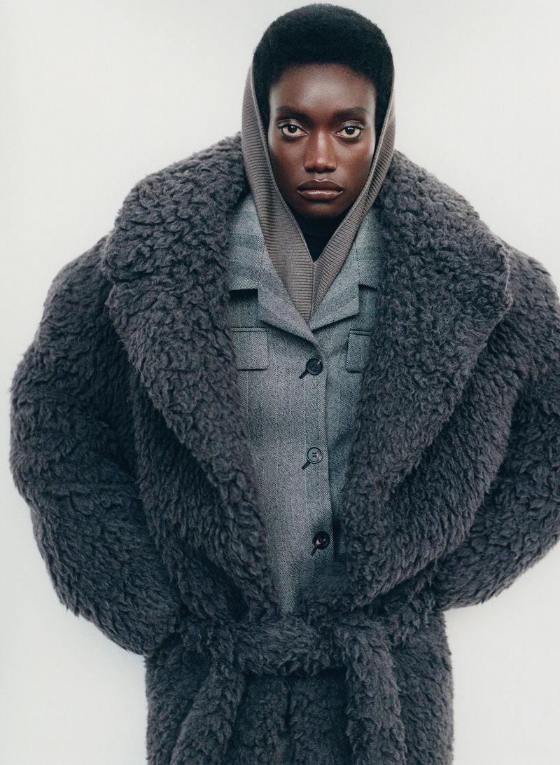 Editorial fashion shot of an adult woman in a gray wool coat and gray suit, looking directly at the camera against a white backdrop.