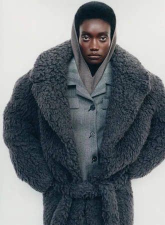 Editorial fashion shot of an adult woman in a gray wool coat and gray suit, looking directly at the camera against a white backdrop.