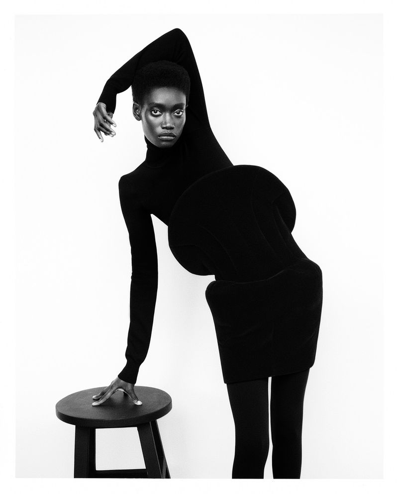 Black and white studio shot of a Black female model in a black turtle neck dress posing with a stool against a white backdrop.