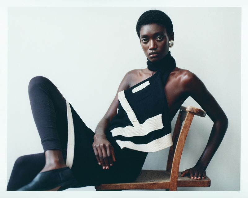 A striking fashion portrait showcases a Black woman seated, adorned in an asymmetrical black and white top, and dark leggings.