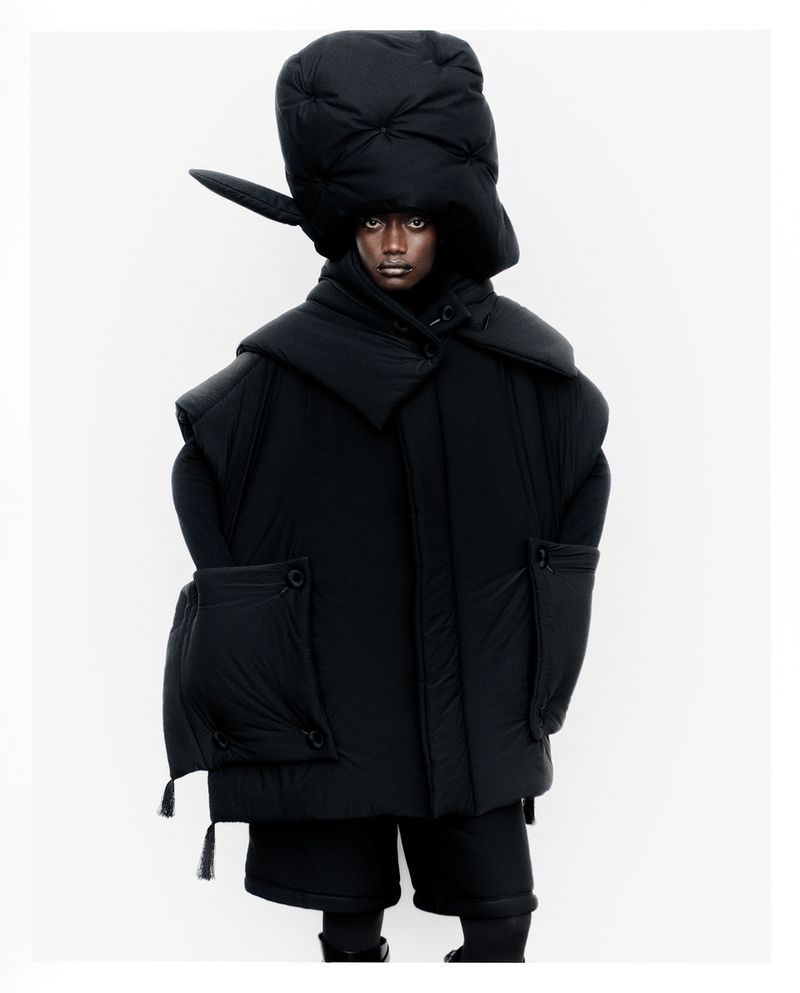 Fashion shot of a Black female model in an oversized black puffer jacket and hat against a white backdrop in a studio environment.