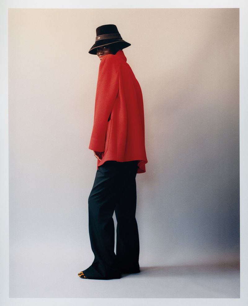 An African American woman in a red jacket and black hat strikes a pose in a fashion shot, set against a neutral studio backdrop.