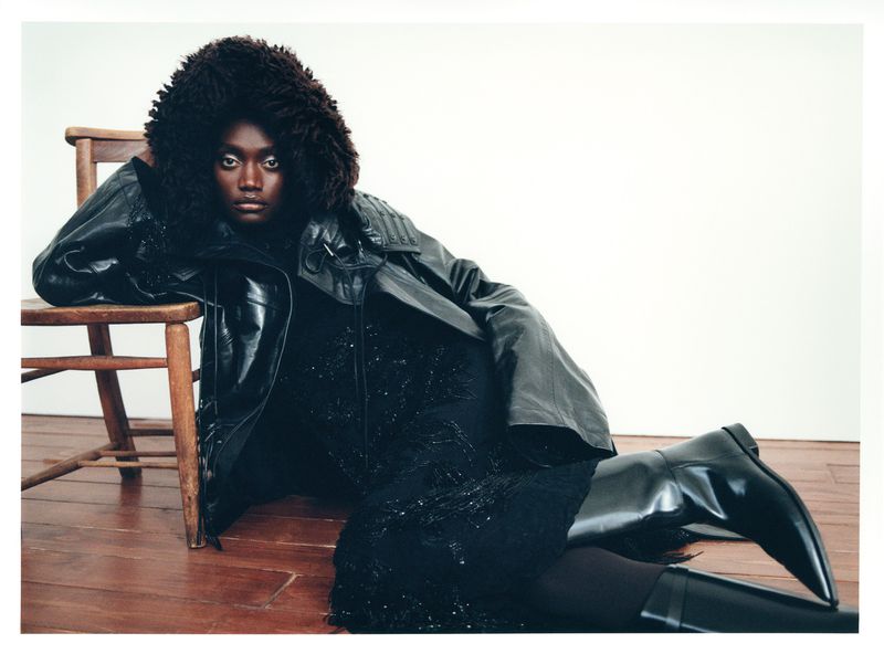 An adult woman poses indoors in an all-black outfit featuring a fur-trimmed hood, leather jacket, dress, tights, and knee-high boots against a neutral backdrop.