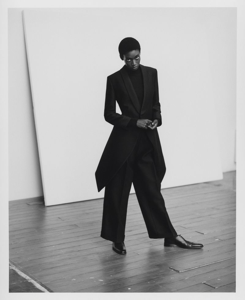 Fashionable woman in a black coat and wide-leg pants strikes a pose in a minimalist photo studio, creating a sleek editorial image.