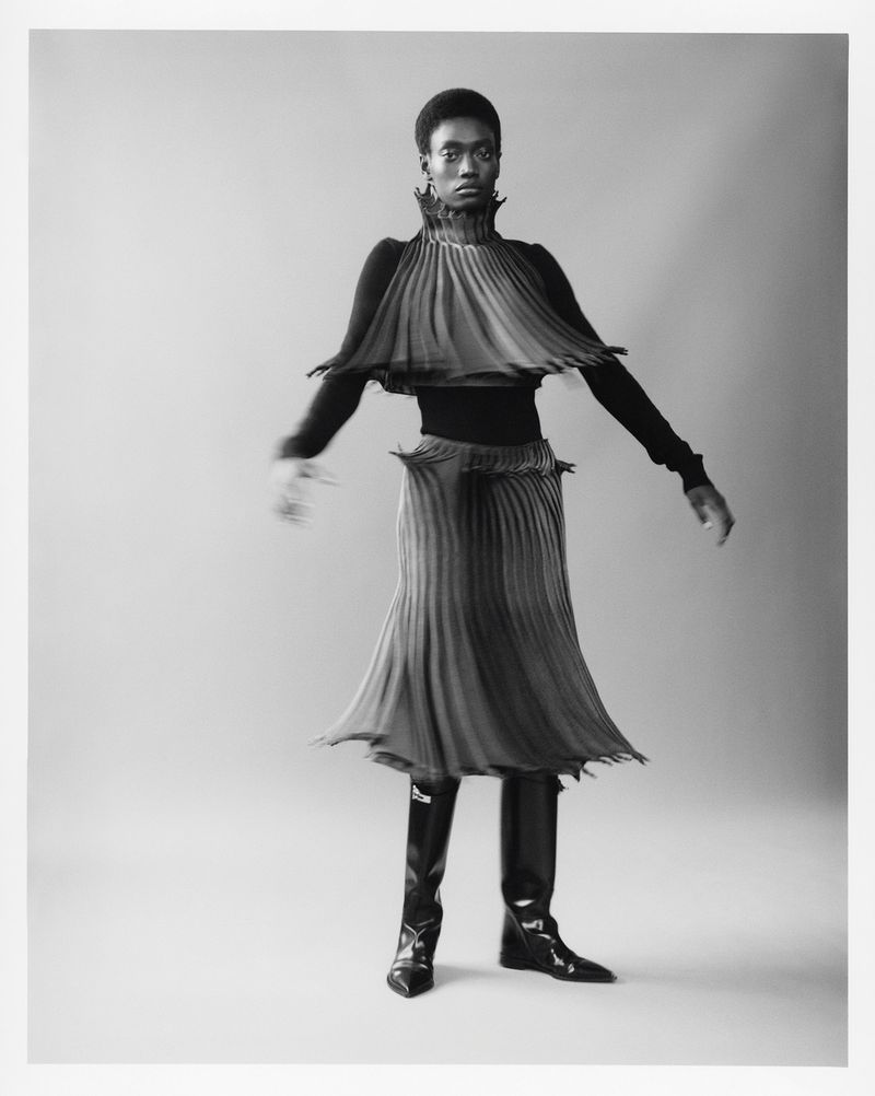 Monochrome fashion shot features African-American model in pleated top and skirt with black boots against a neutral backdrop.
