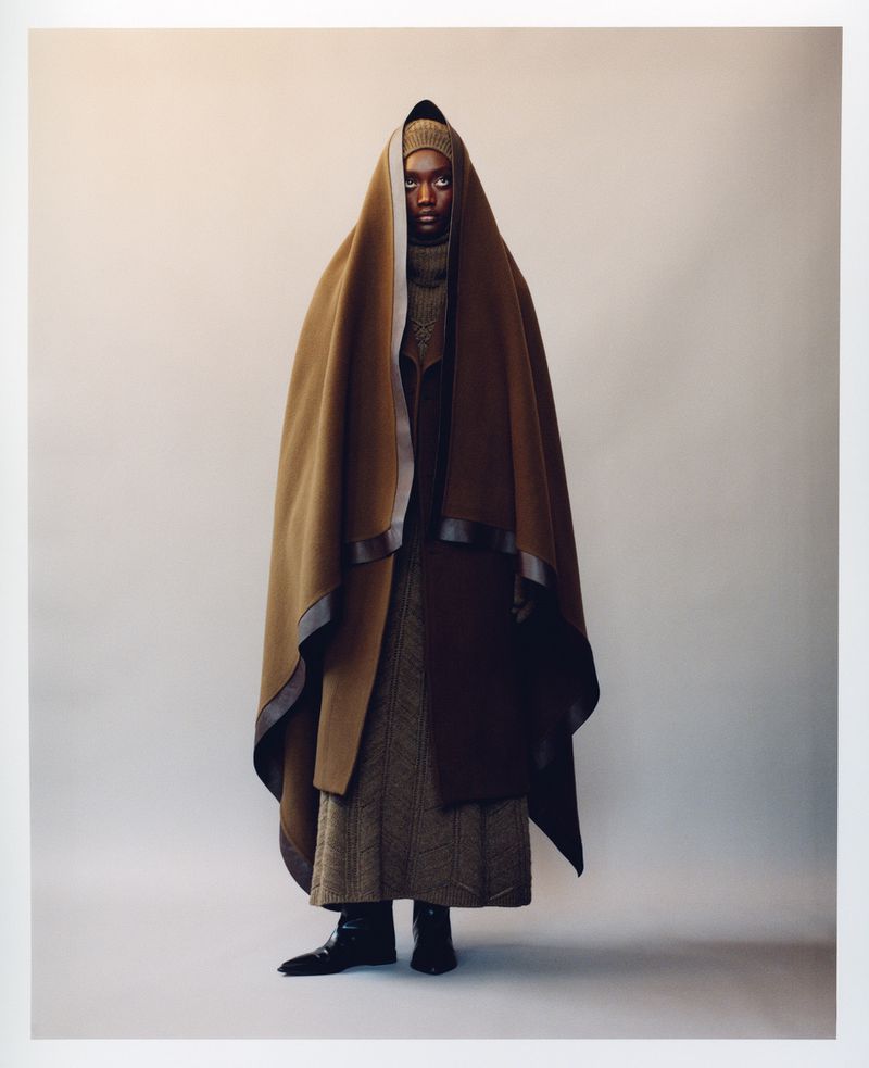Fashion shot of an adult female wearing a draped brown hooded cape, long brown dress, and black boots in a light grey studio setting.