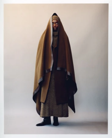 Fashion shot of an adult female wearing a draped brown hooded cape, long brown dress, and black boots in a light grey studio setting.
