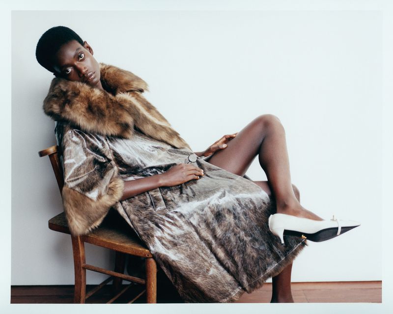 Fashionable woman poses in silver coat with fur collar, sitting on a wooden chair against a white background, wearing white pumps.