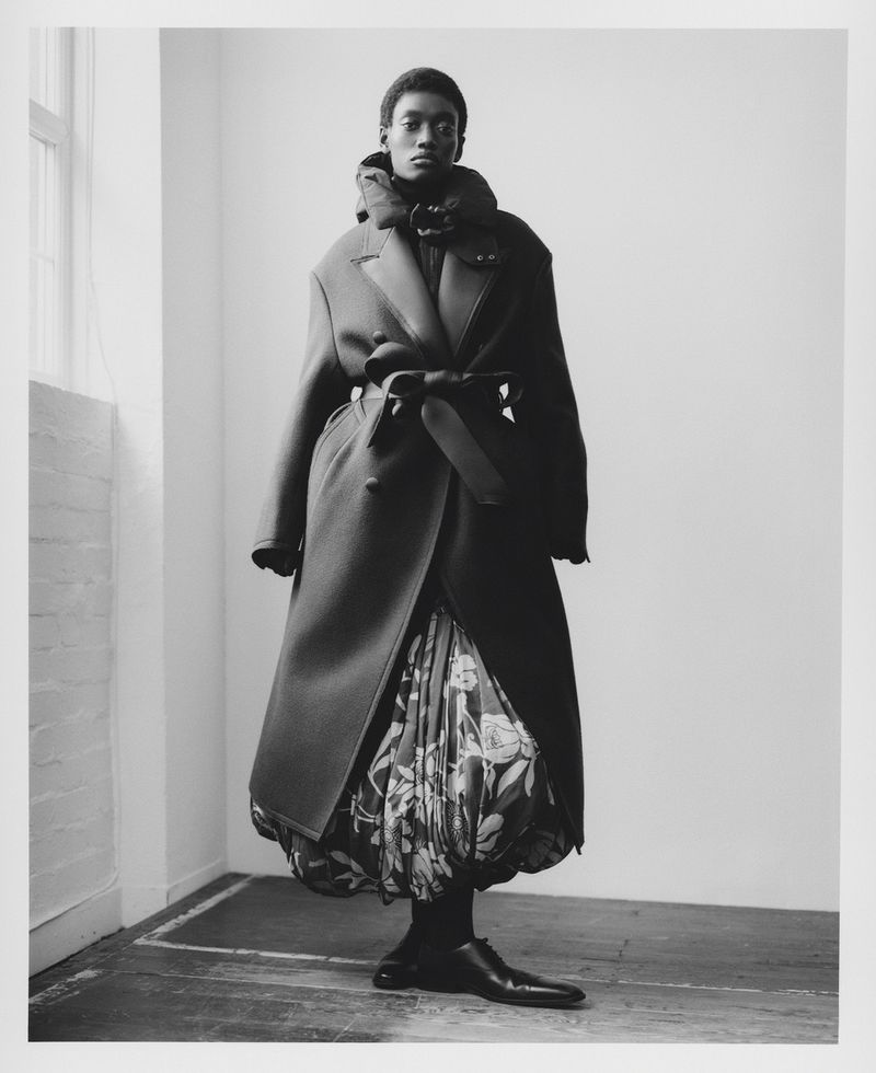 Black-and-white full-body shot of an androgynous fashion model with dark skin wearing a long coat, floral skirt, and leather shoes.