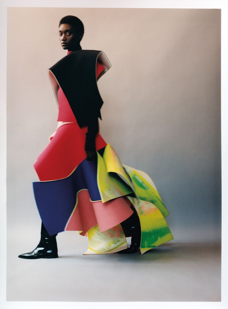Fashion editorial shot: Black model in a sculptural, color-blocked dress against a light gray background in studio lighting.