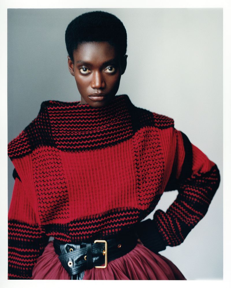 Close-up portrait of a Black woman wearing a red and black textured knit sweater with a wide black belt against a grey background.