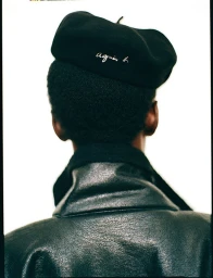 Close-up back view of a model wearing a black agnès b. beret and a textured black leather jacket against a white background.
