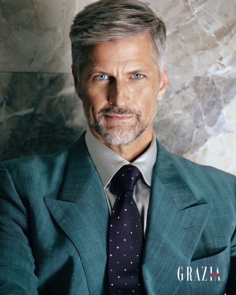 Portrait of an adult male with gray hair wearing a suit and tie, posing in front of a marble wall, with the Grazia logo at the bottom.