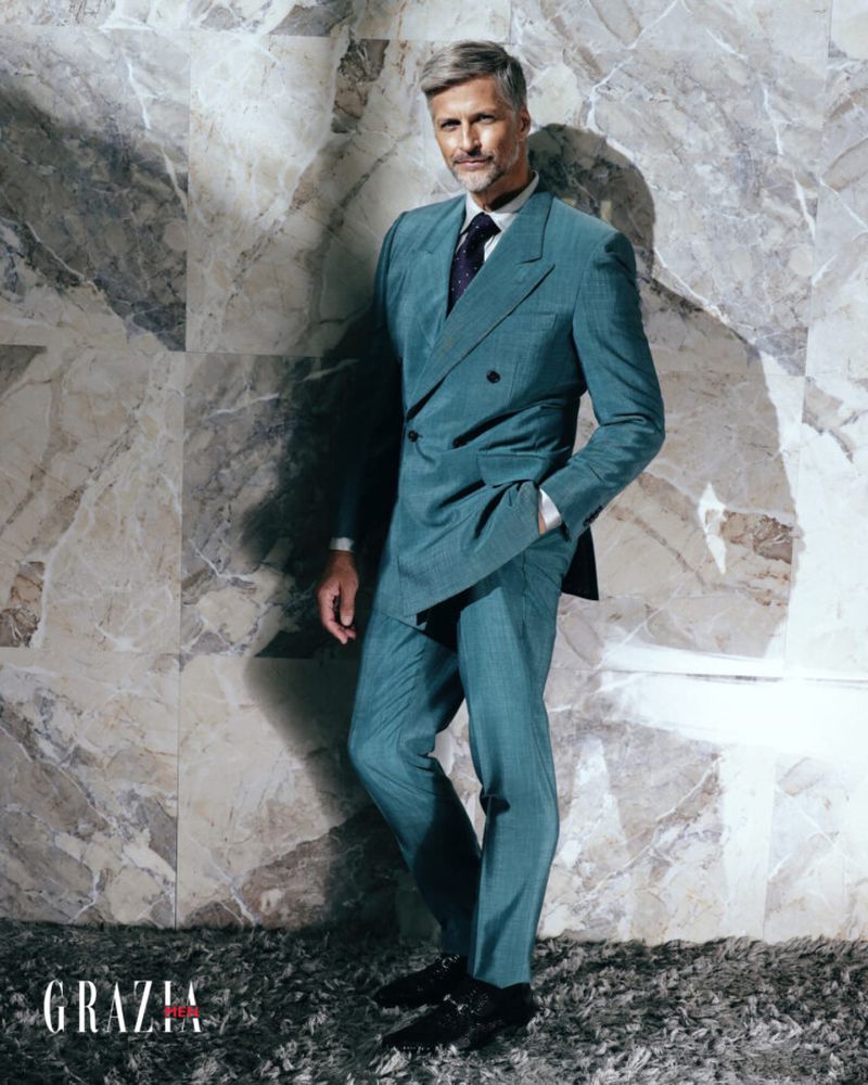 Full-length portrait of a mature adult male with gray hair posing in a double-breasted suit with marble wall and gray floor.