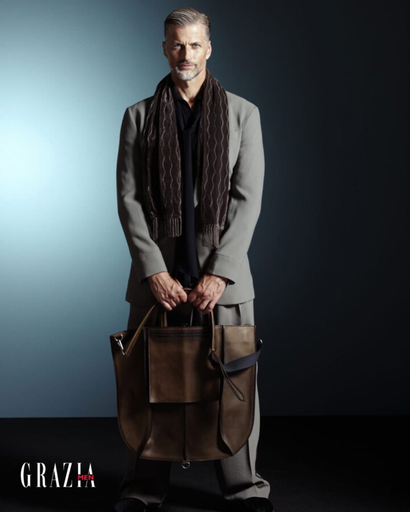 Stylish older male model in tailored suit and scarf, holding a leather bag against a gradient blue background in a studio shot.