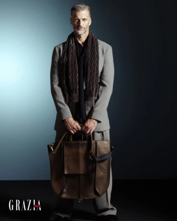 Stylish older male model in tailored suit and scarf, holding a leather bag against a gradient blue background in a studio shot.
