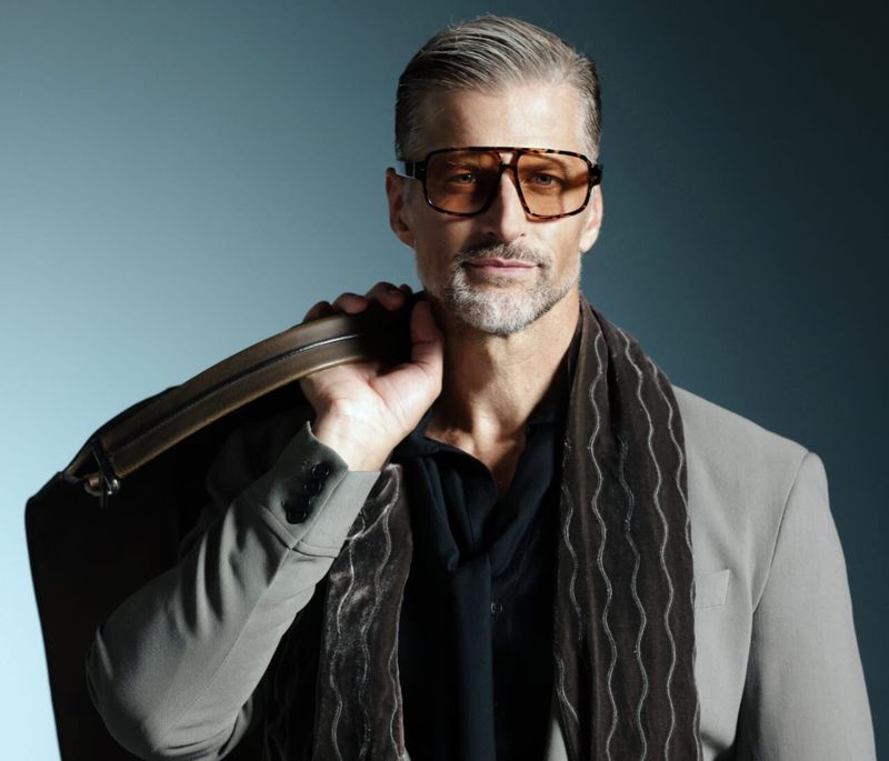 Stylish older man in glasses and blazer poses with a large bag, showcasing fashion and grooming against a muted grey-blue backdrop.