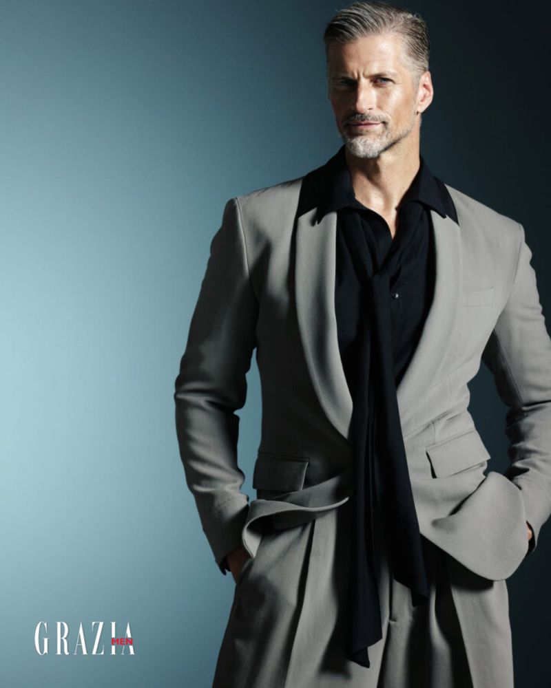Portrait of an adult male with gray hair wearing a gray suit, black shirt and tie against a gradient blue background in a studio setting.