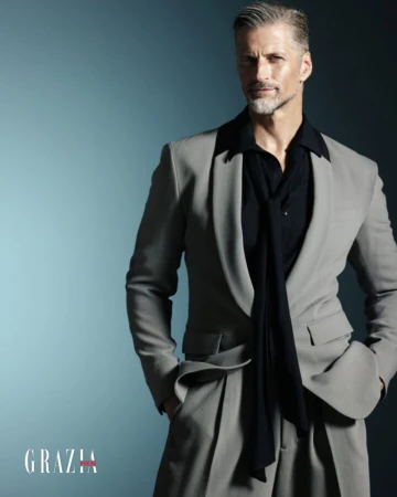 Portrait of an adult male with gray hair wearing a gray suit, black shirt and tie against a gradient blue background in a studio setting.