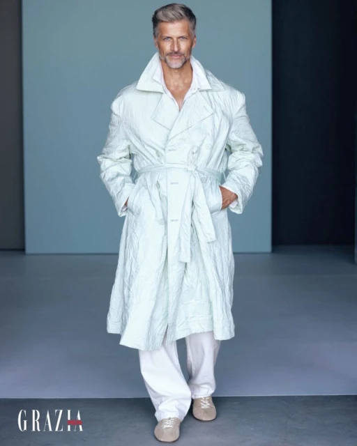 Mature man with gray hair in a light green double-breasted trench coat, walking toward the camera, in a minimalist photo studio.