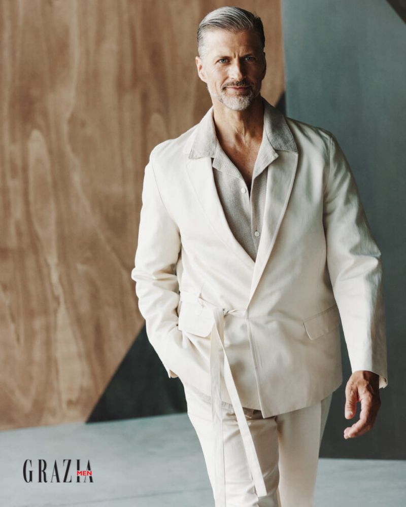 Stylish mature man in a light suit poses indoors against a geometric backdrop, showcasing a blend of modern fashion and classic tailoring.