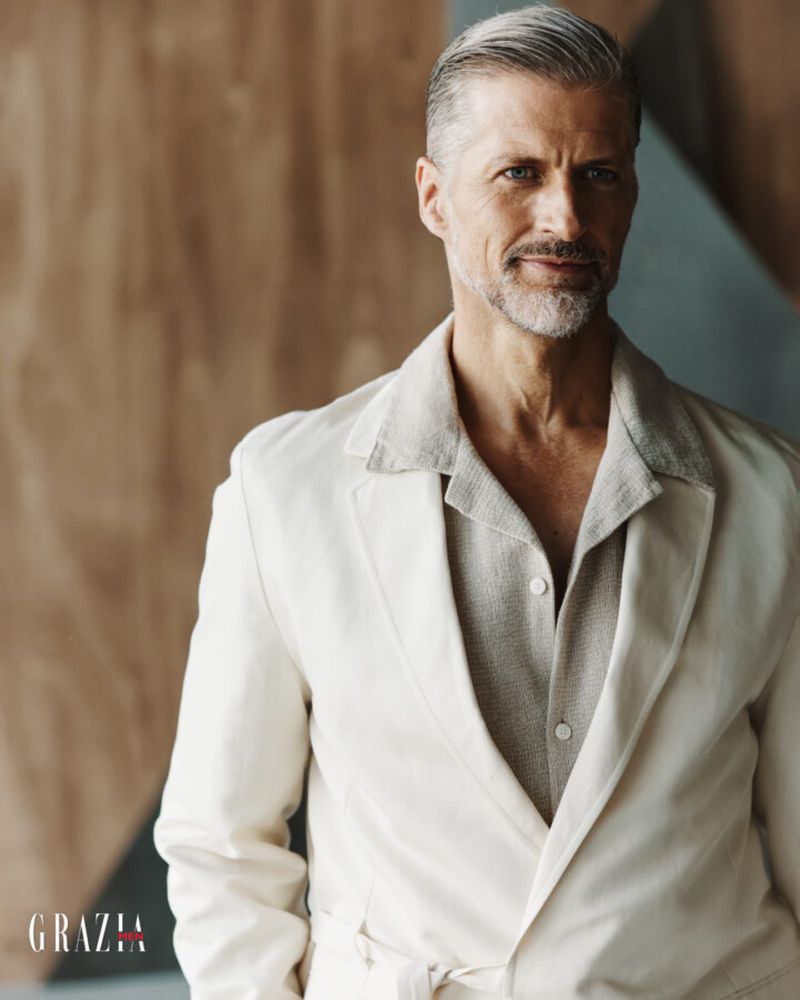 Mature man in a light-colored suit stands indoors against a modern geometric background, captured in a fashion-style portrait.