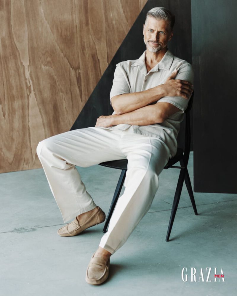 Handsome mature man with gray hair seated on a black chair in a photo studio, wearing a linen shirt and white pants, with warm, soft lighting.